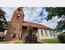 Backsteinkirche mit Satteldach und einem hohen Turm mit Uhr, flankiert von Bäumen; im Vordergrund eine aus Feldsteinen gebaute Skulptur. Der Himmel ist leicht bewölkt, das Gelände besteht aus Rasenflächen und einem gepflasterten Weg.