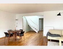 Wohnzimmer mit Holzboden, einem runden Holztisch und vier Stühlen, einer hellen Couch, einer Stehlampe und einer schlichten weißen Wendeltreppe im Hintergrund. Die Wände sind weiß, und eine Tür führt in einen angrenzenden Raum.