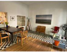 Ein kleiner, schlicht eingerichteter Raum mit Holzboden und schwarz-weiß kariertem Fliesenbereich. Im Vordergrund steht ein dunkler Holztisch mit mehreren unterschiedlichen Stühlen. An der rechten Wand befinden sich ein roter Stuhl, ein großer Ventilator und ein Stativ mit einer Lampe. An der Wand hängen ein Landschaftsbild und ein gerahmtes Porträtfoto.