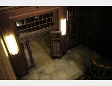 Innenansicht eines Treppenhauses mit dunklem Holzvertäfelungen und gläsernen Türen mit Gitterrahmen. Der Boden ist aus Steinfliesen, und vertikale Wandlampen beleuchten den Raum.