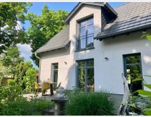 Modernes Einfamilienhaus mit weißer Fassade, großen Fenstern und grauem Schrägdach. Im Vordergrund ein Garten mit üppigem Grün, Terrasse mit Tisch und gelben Stühlen sowie dekorativen Elementen wie einer kleinen Skulptur auf einem Holzstamm.