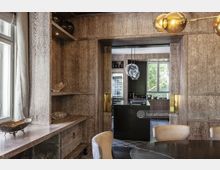 Innenraum eines modernen Wohnbereichs mit Holzvertäfelung und integrierten Regalen an den Wänden, einem Esstisch mit beigen Stühlen im Vordergrund und einer offenen Küche im Hintergrund. Die Küche verfügt über eine zentrale schwarze Insel, hängende Glasleuchten und Fenster, die Blick ins Grüne bieten.