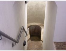 Blick auf eine schmale, gemauerte Wendeltreppe mit weißen Wänden und einem Metallgeländer, die zu einem kleinen, eingelassenen Wasserbecken in einem gewölbten Raum führt.