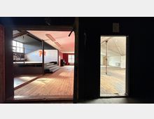 Blick durch zwei Öffnungen in eine ehemalige Veranstaltungshalle mit breitem Holzdielenboden, einer kleinen Bühne mit Stufen und großen Fenstern an der Stirnseite. Rechts ist ein angrenzender leerer Raum mit sichtbaren Stützpfeilern und hoher, schräger Decke zu sehen.