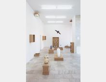 Ein minimalistischer Ausstellungsraum mit weißen Wänden und hellen Fliesenböden, in dem Kunstwerke auf braunen Pappkarton-Sockeln präsentiert werden. Zu den Exponaten gehören Skulpturen, darunter ein Tierkopf und ein bemaltes Schwein, sowie eine schwebende Vogelfigur an der Wand.