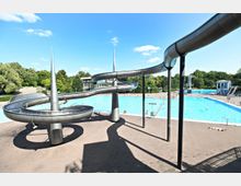 Ein großes Freibad mit einem weitläufigen Schwimmbecken, mehreren Röhrenrutschen aus Metall und einem Sprungturm im Hintergrund. Umgeben von Bäumen und einer gepflasterten Liegefläche, bei klarem Himmel und sonnigem Wetter.