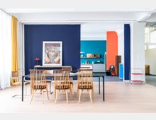 Ein heller Raum mit Holzboden, in dem ein Esstisch mit Holzkreuzstühlen im Vordergrund steht. Die Wände sind in Blau, Orange und Türkis gestrichen; ein Kunstwerk ist an einer blauen Wand angebracht, und im Hintergrund befindet sich eine kleine Küchenzeile mit Regalen und Haushaltsgegenständen.