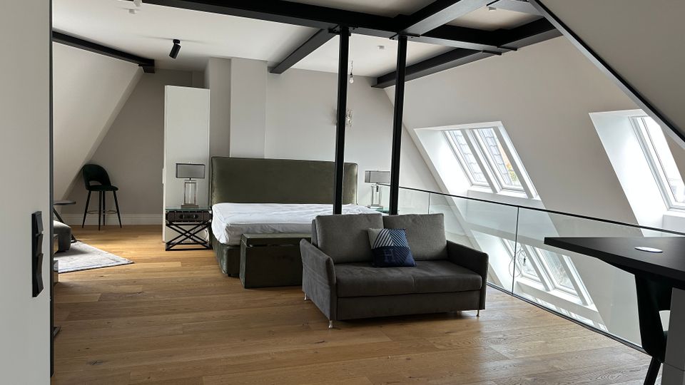 Modernes Dachgeschoss-Schlafzimmer mit sichtbaren schwarzen Stahlträgern, Holzfußboden und schrägen Deckenflächen. Ein Bett und ein Sofa stehen neben einer Glasbrüstung; mehrere Dachfenster sorgen für Tageslicht.