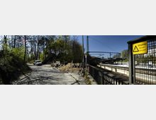 Pflasterstraße neben einer Baustelle mit Sandhaufen und abgesperrtem Bereich, umgeben von Bäumen. Rechts ein Bahnhofsbereich mit Bahnsteig, Gleisen und Warnschild für Hochspannung, im Hintergrund eine Überdachung und Oberleitungen.