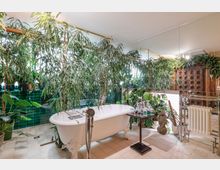 Badezimmer mit freistehender, weißer Badewanne auf Löwenfüßen, umgeben von zahlreichen großen Zimmerpflanzen. Die Gestaltung umfasst grüne Wandfliesen, eine Holzvertäfelung sowie Spiegel, die den Raum optisch erweitern.