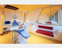 Ein moderner Zahnarzt-Behandlungsraum mit einer blauen Behandlungsstuhl-Einheit, gelbem Boden, weißen Wänden und roten Sitzbänken. Große Spiegel und Fenster schaffen ein helles, offenes Ambiente.