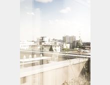 Blick von einer erhöhten Terrasse auf eine urbane Stadtlandschaft mit modernen Wohn- und Bürogebäuden. Auf der Terrasse stehen ein weißer Tisch mit vier Laternen und ein passender Stuhl; im Vordergrund sind einige Pflanzen sichtbar, darüber ein heller Himmel mit wenigen Wolken.