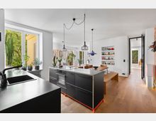 Moderne Wohnküche mit offenem Grundriss, schwarzen Kücheninseln, Holzfußboden und großen Fenstern, die viel Tageslicht einlassen. Im Hintergrund befinden sich ein Essbereich mit Holztisch und Wandschränken sowie eine Glastür, die zu einem weiteren Raum führt.