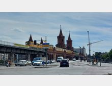 Straßenszene mit der Oberbaumbrücke in Berlin, einer roten Backsteinbrücke mit gotischen Türmen, auf der eine gelbe U-Bahn fährt. Im Vordergrund sind Autos, Motorräder und Straßenverkehrsschilder zu sehen, begleitet von Fußgängern und einem Baukran im Hintergrund.