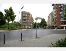 Straßenkreuzung mit Kopfsteinpflaster am Lohmühlenplatz, umgeben von Bäumen und mehrstöckigen Wohnhäusern aus rotem und gelbem Backstein. Im Vordergrund Gehwege und eine Straßenschildmarkierung; parkende Autos und eine begrünte Fläche sind sichtbar.