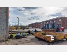Industriehof mit Backsteingebäuden im Hintergrund, Schienenfahrzeugteilen und Maschinen im Vordergrund, darunter ein gelber Transportwagen und ein Gabelstapler. Der Himmel ist teils bewölkt, und im Hintergrund sind Bäume sichtbar.