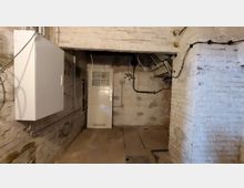 Ein Kellerraum mit weiß gestrichenen Ziegelwänden, sichtbaren Rohren und Kabeln. Im Hintergrund befindet sich eine weiße Tür mit Fenstereinsatz, rechts und links sind Wände mit sichtbaren Abnutzungsspuren.