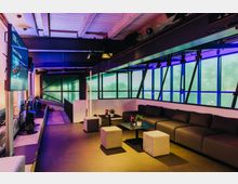 Innenraum einer modernen Lounge mit einem industriellen Design. Große Panoramafenster durchfluten den Raum mit farbigem Licht, während graue Sofas, quadratische Hocker und kleine Tische mit Pflanzen eine einladende Sitzgelegenheit bieten.