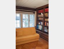 Gemütlicher Raum mit Holzwänden und -decke, einer großen Fensterfront mit weißen Rahmen, gelbem Sofa und eingebautem Bücherregal rechts, das mit Büchern und Ordnern bestückt ist. Der Boden besteht aus glänzendem Holzparkett.