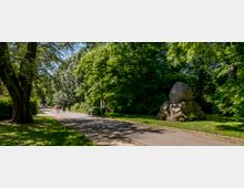 Ein gepflegter Park mit einer asphaltierten Gehwegallee, umgeben von großen, dicht belaubten Bäumen. Rechts des Weges stehen mehrere große Felsbrocken, und entlang des Weges befinden sich Bänke sowie eine Laterne.