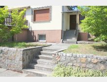 Eingangsbereich eines Einfamilienhauses mit gelber Fassade und braunem Sockel. Mehrere Steintreppen führen durch einen kleinen, von Grünflächen und Ziermauern eingerahmten Vorgarten zur Haustür.