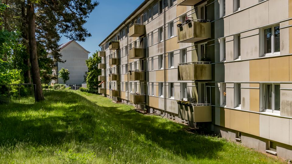 Langer, mehrstöckiger Wohnblock mit beigen Fassadenelementen, vielen Fenstern und vorspringenden Balkonen entlang einer schmalen Grünfläche. Links säumen hohe Bäume und dichtes Gebüsch den Rasenstreifen; im Hintergrund ist ein weiteres Gebäude zu sehen.
