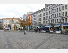 Pflastersteinplatz in einer Stadt mit umliegenden Gebäuden im Stil des europäischen Altbaus, Straßengeschäften, einem U-Bahn-Eingang, einer Statue auf einem Sockel und vereinzelten Sitzbänken entlang des Platzes. Im Hintergrund stehen Bäume mit herbstlichem Laub.