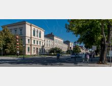 Straßenansicht mit einem historischen, sandfarbenen Gebäude im klassizistischen Stil, das von Bäumen und Fußgängern umgeben ist. Der Platz zeigt verschiedene Straßenelemente wie Laternen, parkende Fahrräder und Autos sowie Passanten unter blauem Himmel.