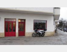 Straßenszene mit einem Gebäude, das ein Geschäft mit der Aufschrift "Location Networx" zeigt, erkennbar durch rote Türen und Fensterrahmen. Vor dem Geschäft steht ein Motorrad auf einem Gehweg neben einer Reihe parkender Fahrräder.