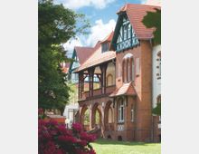 Backsteingebäude im historischen Stil mit Fachwerkgiebeln, gewölbten Arkaden und einer großen Holzveranda. Im Vordergrund blühende Rhododendren und grüne Rasenflächen; im Hintergrund blauer Himmel und Bäume.