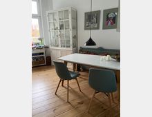 Essbereich in einem hellen Raum mit Holzfußboden, einem weißen Esstisch und zwei gepolsterten Stühlen. Im Hintergrund stehen ein weißer Vitrinenschrank, eine Sitzbank mit bunten Kissen und an der Wand hängen zwei gerahmte Fotos.