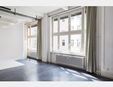 Heller Raum mit großen Fenstern, die viel Tageslicht hereinlassen. Der Boden ist dunkel, an den Heizkörpern unter den Fenstern hängen lange, helle Vorhänge, und eine weiße, gebogene Fläche befindet sich links im Bild, möglicherweise Teil eines Fotostudios.