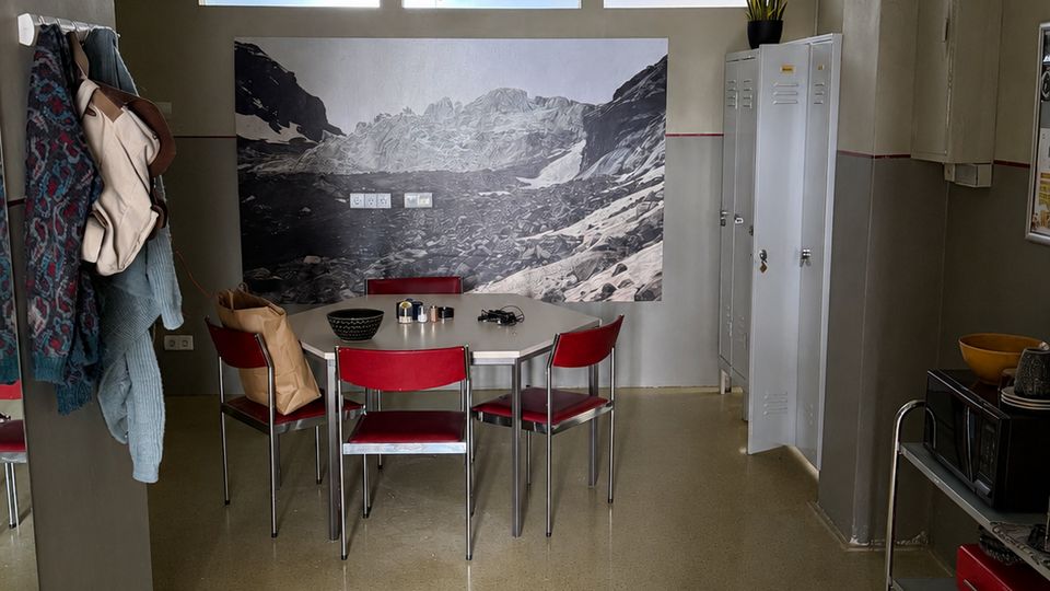 Innenraum eines schlichten Aufenthalts- oder Pausenraums mit rundem Tisch und roten Stühlen vor einer großen Fotowand mit Berglandschaft. Hohe Fenster mit Rollos, graue Wände, ein weißer Spind und ein Servierwagen mit Küchenutensilien prägen den Raum.