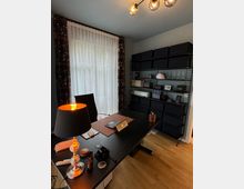 Moderner Arbeitsplatz in einem Raum mit hellem Holzboden. Ein Schreibtisch mit Laptop, Tischlampe und Bürobedarf steht vor einem großen Fenster mit weißen Vorhängen und gemusterten Seitenschals. An der Wand befindet sich ein Regal mit schwarzen Aufbewahrungsboxen und Dekorationsgegenständen.