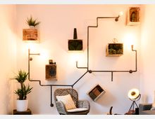 Wohnzimmer mit modernem Wanddesign, bestehend aus Rohrleitungen, die als Rahmen für Holzkisten mit Dekor wie Pflanzen und Büchern dienen. Im Vordergrund steht ein geflochtener Sessel mit Kissen, daneben eine Topfpflanze und eine Stehlampe.