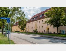 Straßenansicht mit mehrstöckigen Wohngebäuden in beige, roten Satteldächern und kleinen Dachfenstern, umgeben von Bäumen und Rasenflächen. Ein Straßenschild mit der Aufschrift „Clara-Zetkin-Str.“ steht links neben einem Gehweg.