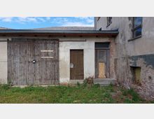 Eine alte Gebäudefassade mit einer großen Holztür, einer kleineren Holztür und einem zugemauerten Eingang. Die Wand besteht aus Putz und teilweise freigelegten Ziegeln, der Boden ist von Gras und Pflanzen bewachsen.