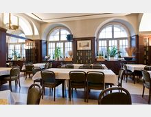 Ein Speisesaal mit hohen Decken, großen Rundbogenfenstern und gedeckten Tischen mit weißen Tischdecken. Die Wände sind mit dunklem Holz verkleidet, und auf Fensterbänken stehen Pflanzen und Dekorationsobjekte.