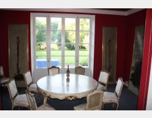 Ein eleganter Raum mit roten Wänden und großen Fenstern, die auf einen grünen Garten blicken. Im Vordergrund steht ein runder Holztisch mit geschnitzten Stühlen, und an den Wänden hängen schmale, gerahmte Wandbilder.