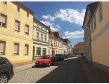 Eine gepflasterte Straße in einer Altstadt mit mehrstöckigen, hellen Gebäuden aus verputztem Mauerwerk, eingerahmt von geparkten Autos. Im Hintergrund sind rote Ziegeldächer und ein sonniger Himmel mit wenigen Wolken zu sehen.