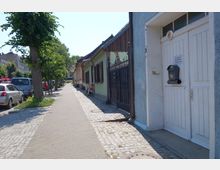 Eine gepflasterte Gehwegstraße mit parkenden Autos auf der linken Seite und einer Reihe kleiner, bunter Häuser mit Zäunen und Fenstern auf der rechten Seite. Im Vordergrund ist eine große weiße Tür mit einem schwarzen Briefkasten zu sehen.