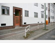 Mehrfamilienhaus mit weißer Fassade und rotem Backsteinsockel; eine Eingangstür mit horizontalen Glasstreifen ist von Ziegelsteinen umrahmt. Vor dem Gebäude verläuft ein Gehweg mit Geländer, neben einem Bürgersteig aus Kopfsteinpflaster.