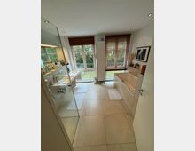 Modernes Badezimmer mit großer, bodengleicher Dusche auf der linken Seite, einem freistehenden Waschbecken und einer Badewanne rechts. Zwei große Fenster mit Holzjalousien bieten Blick ins Grüne, während helle Fliesenbodenmatten und dezente Beleuchtung den Raum ausfüllen.