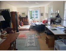 Helles Wohnzimmer mit mehreren Bereichen: links ein Essbereich mit Holztisch und Stühlen, rechts ein Schreibtisch mit Lampen und Büroartikeln, sowie ein kleiner Wohnbereich mit Sofa und Pflanzen vor einem Fenster. Möbel aus Holz und Teppiche schaffen eine gemütliche Atmosphäre.