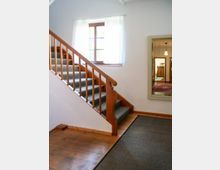 Heller Innenflur mit Holztreppe und Teppichstufen, weiß gestrichenen Wänden und Holzdielenboden. Ein Fenster mit halbtransparenten Vorhängen sowie ein großer Wandspiegel, der einen angrenzenden Raum zeigt, sind zu sehen.