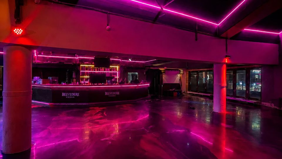 Innenraum eines modernen Nachtclubs mit geschwungener Bar und spiegelglänzendem Boden, der pinke und rote Neonlichter reflektiert. Sichtbare Deckenbalken, LED-Lichtstreifen und Stützsäulen rahmen den offenen Raum mit großen Fensterfronten auf der rechten Seite.