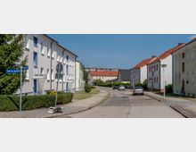 Eine ruhige Wohnstraße mit zweigeschossigen Mehrfamilienhäusern in heller Putzoptik, roten Ziegeldächern und gepflegten Gehwegen. Vereinzelte parkende Autos und kleine Vorgärten mit Hecken säumen die Straße.