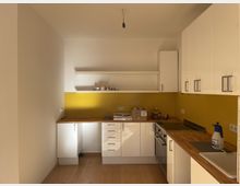 Eine kleine, moderne Einbauküche mit weißen Schränken und gelber Rückwand. Die Arbeitsflächen bestehen aus hellbraunem Holz, und es gibt offene Regale sowie eine Edelstahlspüle und Elektrogeräte wie einen Herd und eine Backofenleiste.