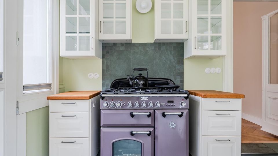 Helle Wohnküche mit großem Gasherd und Backofen in der Mitte, flankiert von weißen Unterschränken mit Holzarbeitsplatten. Darüber hängen weiße Hängeschränke mit Glastüren; die Rückwand ist mit dunkelgrauen Fliesen gestaltet, links ein hohes Fenster und rechts ein Türdurchgang.