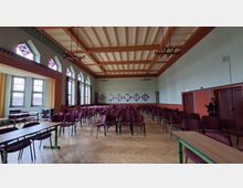 Großer Veranstaltungssaal mit Parkettboden und vielen in Reihen aufgestellten bordeauxfarbenen Stühlen, seitlich ergänzt durch lange Tische. Hohe Fenster mit farbigen Glasfeldern, eine holzverkleidete Kassettendecke mit hängenden Leuchtern und eine Rückwand mit geometrischen Wandtafeln prägen den Raum.